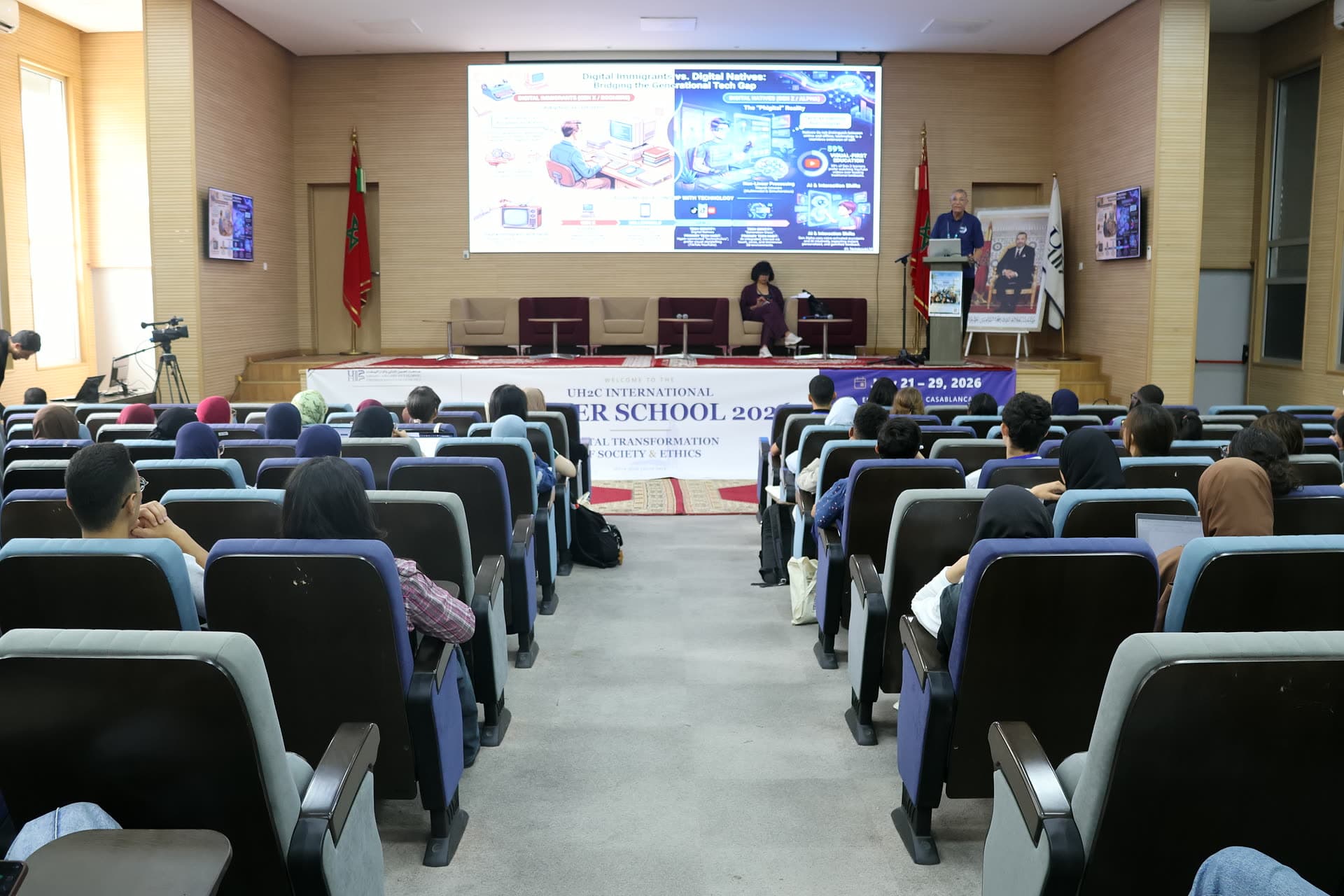 Wide view of the stage during the morning lectures, speaker at the podium with a colourful slide on the big screen.