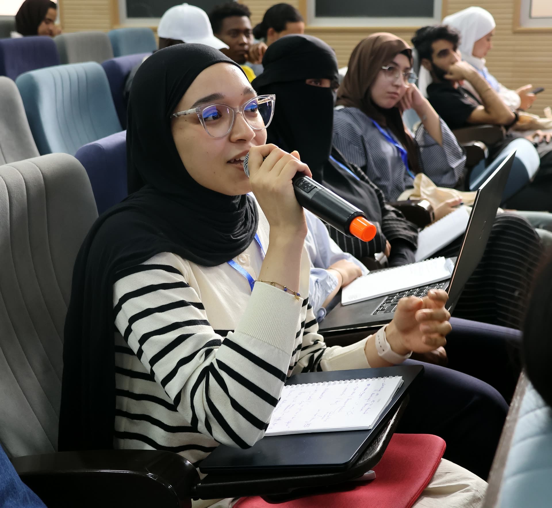 A participant asking a question at the microphone, notebook and laptop on her lap.