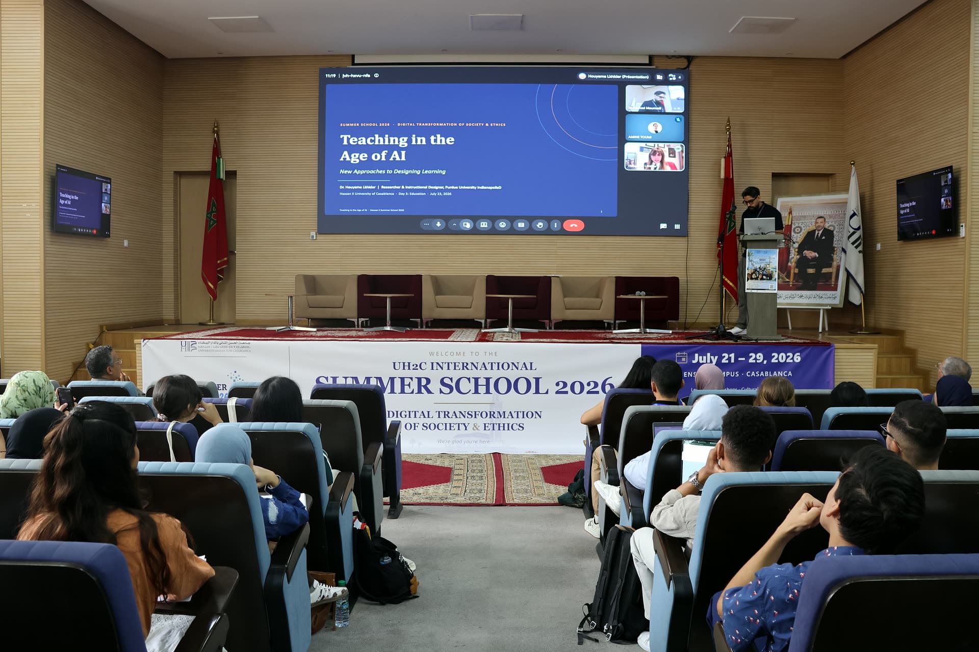 The auditorium’s big screen showing the “Teaching in the Age of AI” online lecture, hosts on stage beside it.
