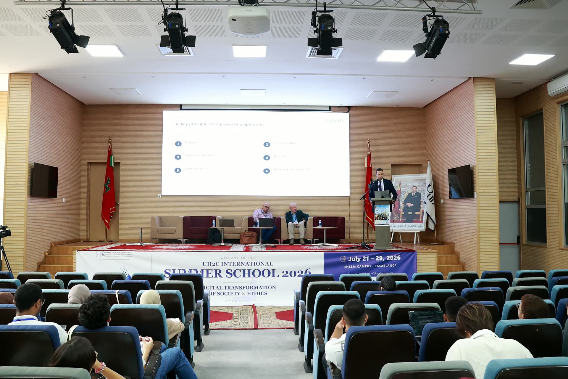 Wide view of the stage during the morning lectures, speaker at the podium with panellists seated.