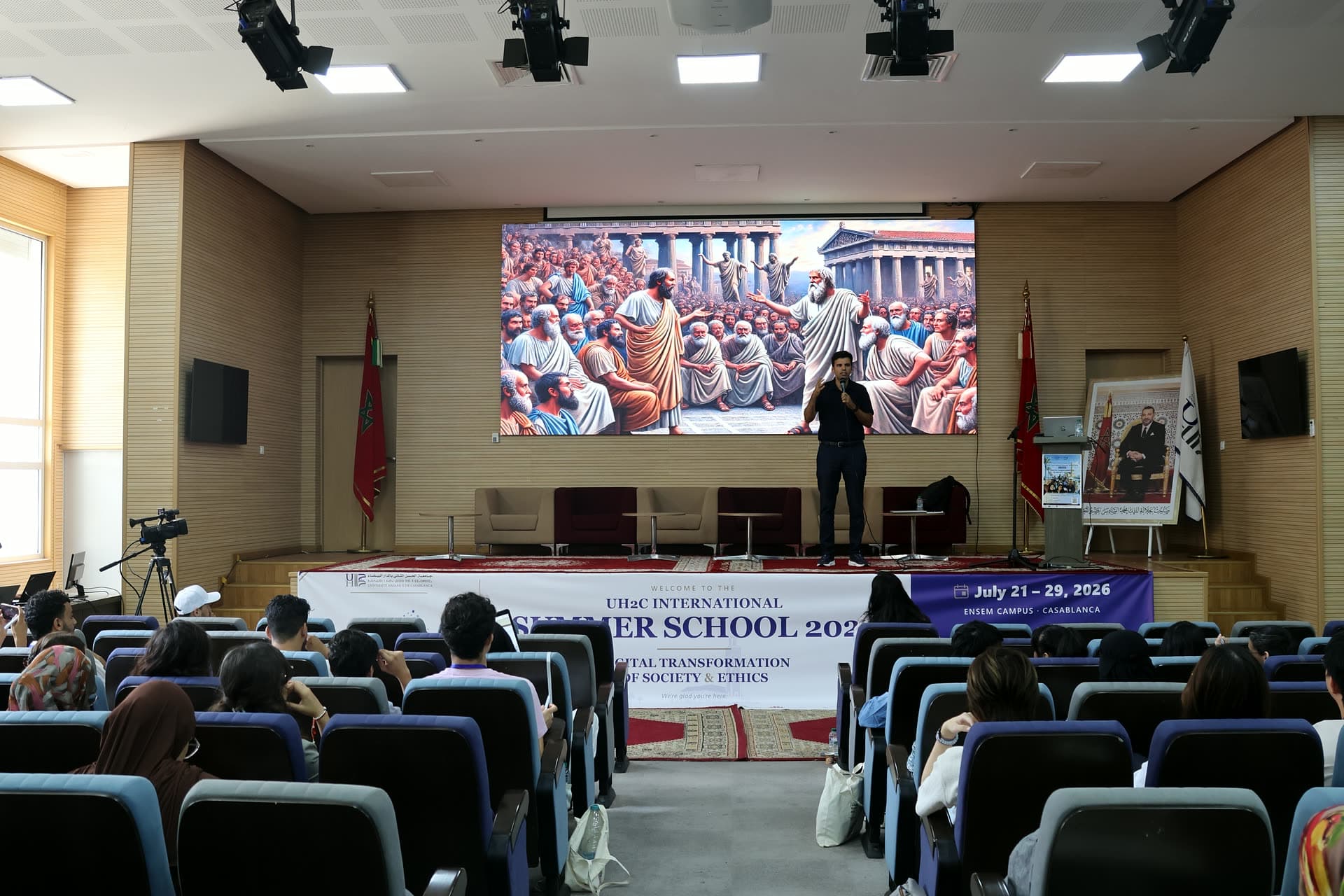 Speaker on stage beneath a giant screen showing a classical agora painting during the responsible-AI lecture.