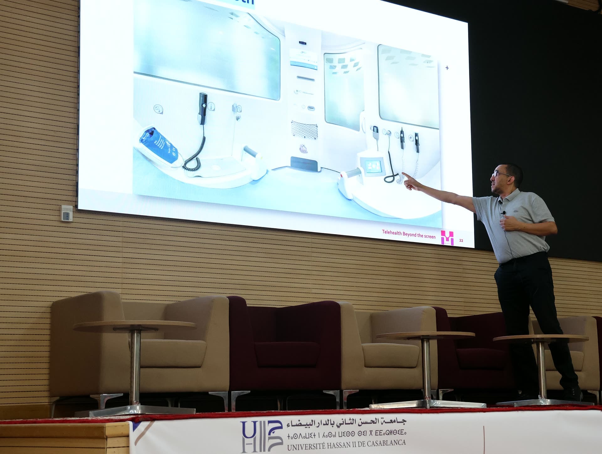 Speaker on stage pointing at a slide showing telehealth stations during the morning lecture.