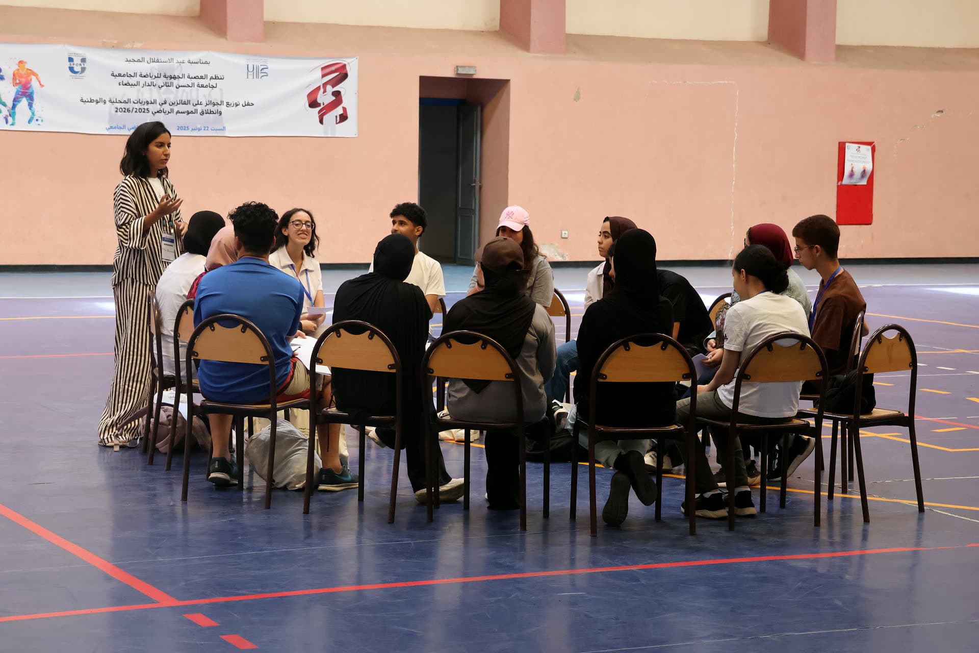 Facilitator leading a circle of participants seated on chairs in the sports hall.