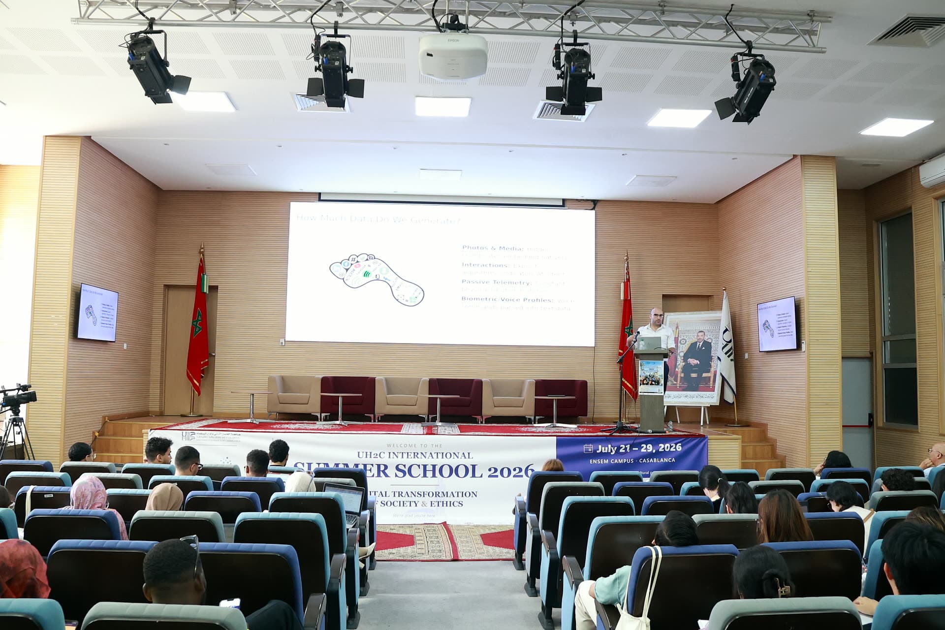 Wide view of the auditorium stage during the morning lectures, a mapping slide on the screen.