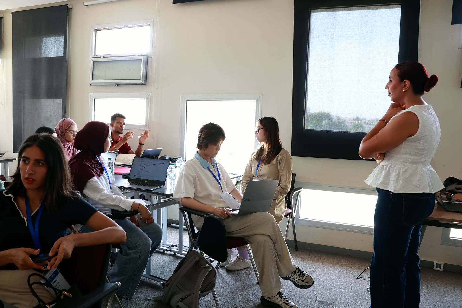Workshop room with teams at laptops and the facilitator standing by the window.
