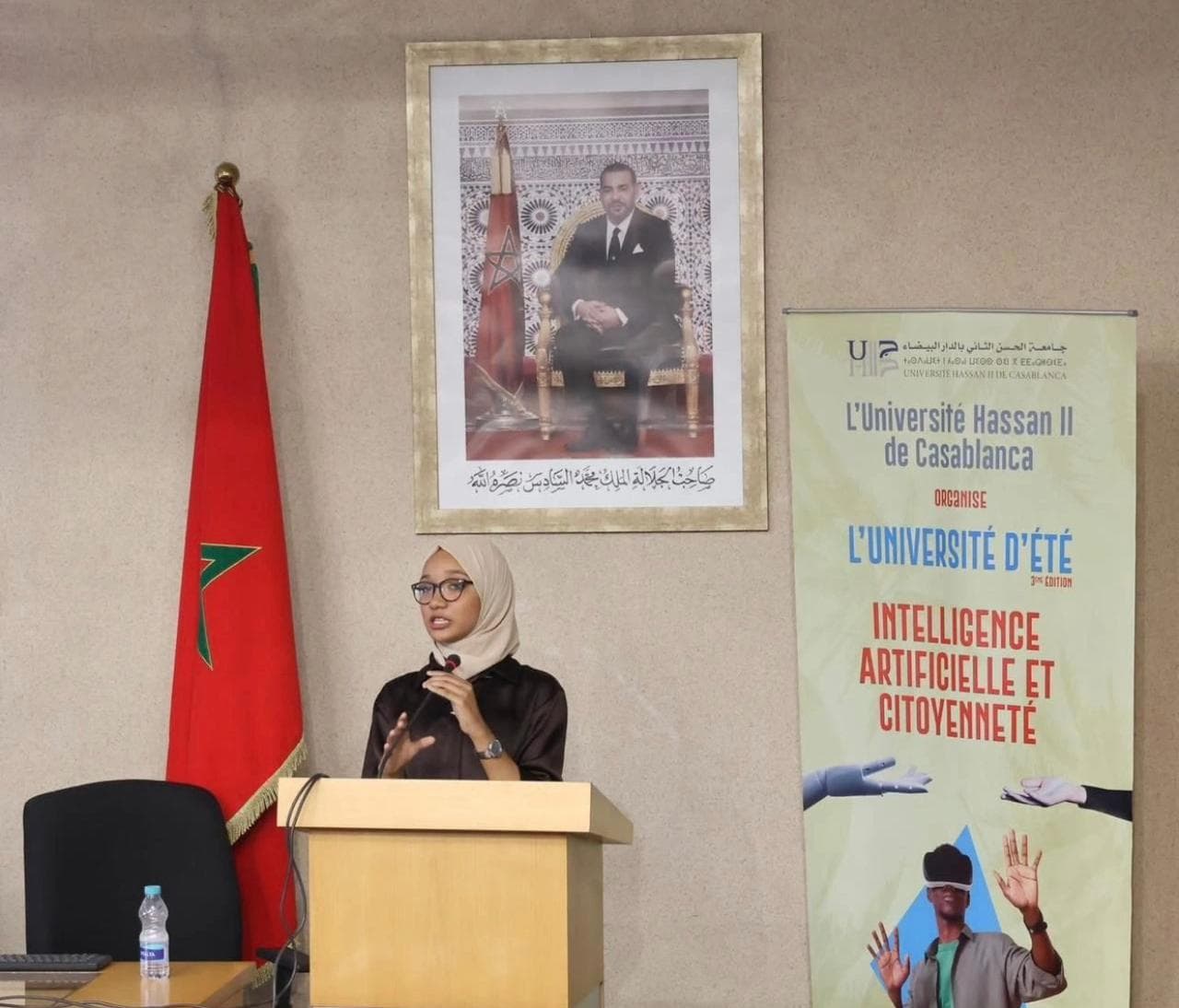 Female speaker delivering a keynote at the podium, beside the Moroccan flag and the official poster.