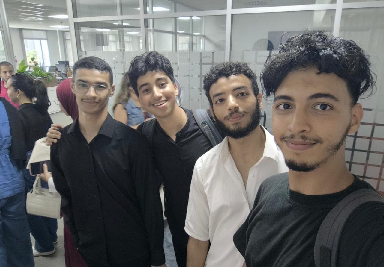 Four students smiling for a selfie in the glass-walled corridor of the library.
